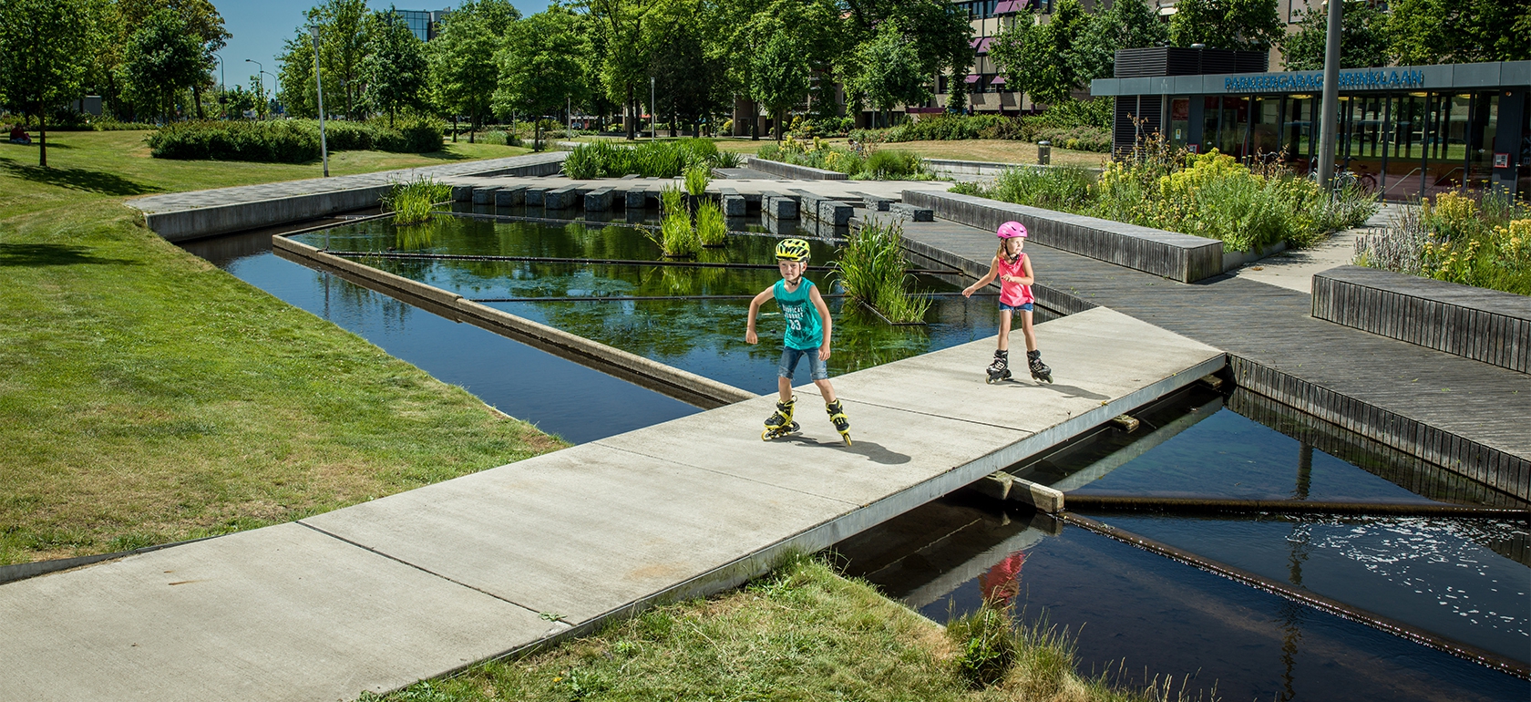 Twee kinderen skeeleren over een pad langs een waterpartij in een groen aangelegd stadspark